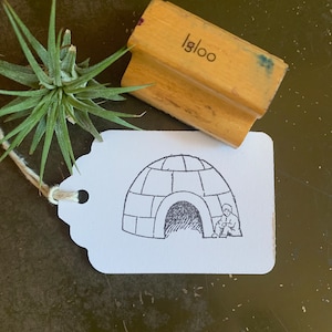 May include: A white tag with a black and white illustration of an igloo with a person inside. The tag has a hole punched in the top left corner and a piece of twine is threaded through the hole. The tag is labeled "igloo" in the top right corner.