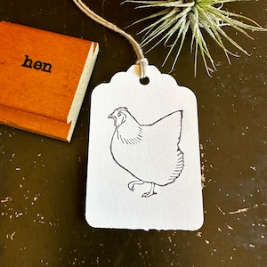 Puede incluir: Una etiqueta de regalo blanca con un dibujo de línea negra de una gallina. La etiqueta está atada con un cordel y tiene un agujero perforado en la parte superior.