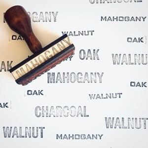 May include: A wooden stamp with the word "MAHOGANY" printed on the stamp face. The stamp is dark brown with a wooden handle. The background is white with the words "MAHOGANY", "OAK", "WALNUT", and "CHARCOAL" printed in a repeating pattern.