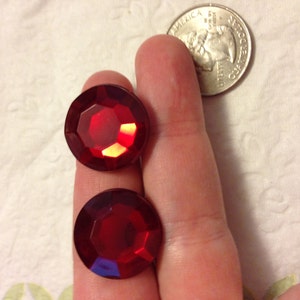 May include: Two red, faceted, round, glass gems. The gems are about the size of a US quarter.