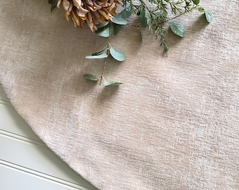 May include: A circular beige fabric mat with a textured, slightly distressed surface finish. A decorative arrangement of a large, dried, light brown flower and green foliage stems rests on the upper edge of the circular textile piece.