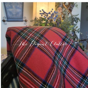 May include: A red fabric featuring a classic tartan plaid pattern with intersecting lines in green, blue, white, and black. The textile is draped, showing the diagonal orientation of the weave. The text The Elegant Clutter is overlaid in white.