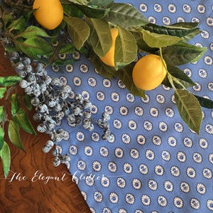 May include: A blue tablecloth with a white floral pattern is adorned with faux lemons and greenery. A sprig of blue berries adds a contrasting color. The Elegant Clutter is written in cursive at the bottom.