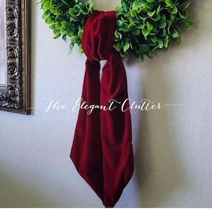 May include: A green wreath with a deep red velvet scarf tied around it. The text "The Elegant Clutter" is written in cursive across the image. The wreath is made of small, dense green leaves.