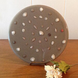 May include: A round, gray decorative plate adorned with various seashells. The plate is displayed on a metal stand and rests on a wooden surface. A single white flower with green leaves lies nearby, adding a touch of natural beauty.