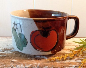May include: A ceramic mug with a brown handle and rim, and a white and brown exterior. The mug features hand-painted illustrations of a tomato and squash. The mug is sitting on a distressed wooden surface.