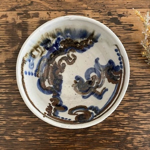 May include: A white ceramic bowl with a blue and brown abstract design. The bowl has a slightly raised rim and a smooth, glazed finish.