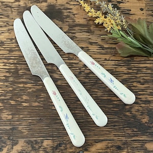 May include: Three silver butter knives with white handles decorated with floral designs in pink, blue, and green.