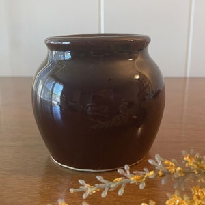 May include: A dark brown ceramic jar with a rounded body and a slightly flared rim. The jar has a glossy finish and a white rim.