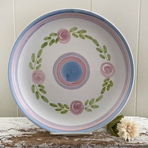 May include: A white ceramic serving bowl with a pink and blue floral design. The bowl features a central circle with concentric rings of pink and blue, surrounded by a wreath of pink roses and green leaves.
