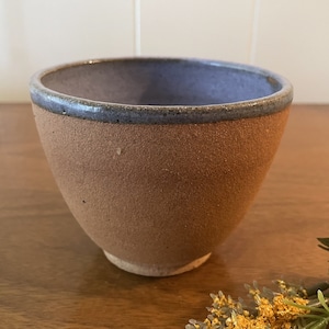 May include: A small, brown ceramic bowl with a blue rim. The bowl has a rough, textured surface.