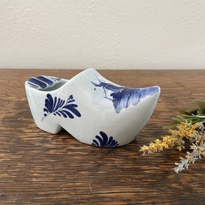 May include: A blue and white ceramic Dutch shoe shaped ashtray with a windmill design. The shoe is sitting on a wooden surface.