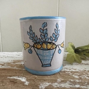 May include: A small, white ceramic mug with a blue rim and a hand-painted design of a blue and yellow flower arrangement in a blue and white patterned vase.