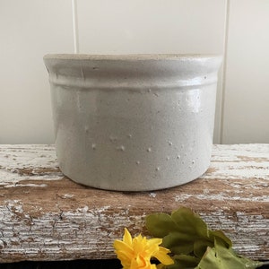 May include: White ceramic crock with a glazed finish. The crock has a rounded shape and a slightly flared rim. It is sitting on a weathered wooden surface.