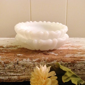 May include: Two white milk glass bowls with a scalloped edge. The bowls are stacked on top of each other and are sitting on a piece of weathered wood.