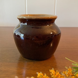 May include: A small, dark brown ceramic pot with a glossy finish. The pot has a rounded body and a slightly flared rim with a lighter brown color. The pot is likely vintage or antique.