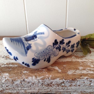 May include: A white ceramic clog with blue floral and windmill designs. The Dutch-style shoe features a detailed painted scene on the side and decorative blue accents. This decorative item is a unique piece of home decor.