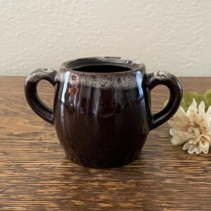 May include: A small, dark brown ceramic sugar bowl with two side handles. The rim features a decorative, textured white and grey drip glaze pattern that contrasts with the smooth, glossy finish of the main body of the rounded vessel.