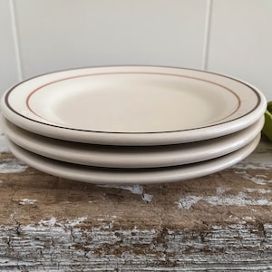May include: A stack of four off-white ceramic plates. Each plate has a thin brown and reddish-brown border. The plates are stacked on a weathered, light brown wooden surface.