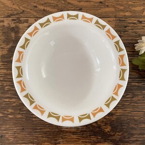 May include: White ceramic bowl with a repeating geometric pattern in shades of orange and olive green around the rim. The bowl is set against a dark wood surface, showcasing a vintage aesthetic. A decorative floral element is visible in the background.