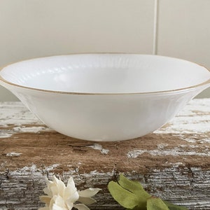 May include: A white ceramic bowl with a gold rim. The bowl has a gently curved shape and sits on a rustic wooden surface. The bowl is empty and appears to be clean. The bowl is likely for serving food.