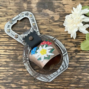 May include: A silver bottle opener with a painted cowbell attached. The cowbell is decorated with colorful flowers and has a brown, metallic finish.