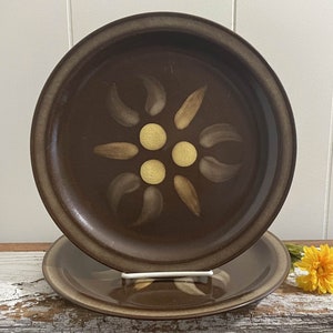 May include: Two brown ceramic dinner plates with a stylized flower design in shades of brown and yellow. The plates have a raised rim.