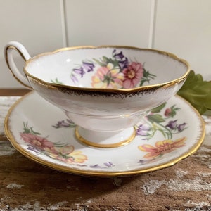May include: A white teacup and saucer set with a floral design in pink, yellow, and purple. The edges are trimmed with gold. The teacup has a curved handle and sits on a pedestal base. The saucer mirrors the teacup's design.