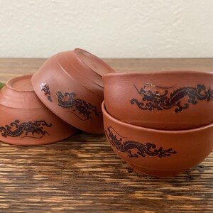May include: Four terracotta teacups with black dragon designs. The cups are stacked on top of each other.