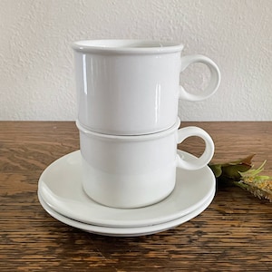 May include: Two white ceramic coffee cups stacked on top of each other with a white saucer underneath. The cups have a simple design and are perfect for everyday use.