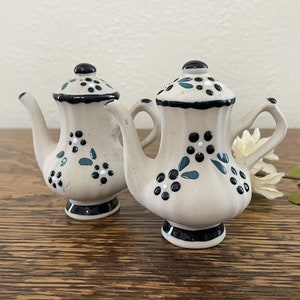 May include: A set of two white ceramic salt and pepper shakers shaped like teapots. The shakers are decorated with black and green floral designs.