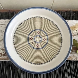 May include: A round ceramic plate featuring a wide blue rim and a central decorative pattern that mimics a coiled basket weave. The design consists of concentric circles in beige, blue, and small maroon cross-shaped accents at the center.