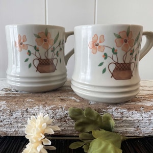 May include: Two white ceramic mugs with a floral design. The mugs feature a basket of orange flowers with green leaves.