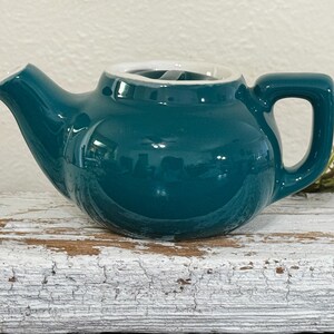 May include: A small, glossy, dark teal ceramic teapot with a rounded body, a short spout, and a matching handle. The teapot features a white interior rim and a lid with a small central knob, showcasing a smooth, reflective finish throughout the exterior.
