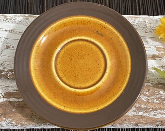May include: A brown and yellow ceramic saucer with a raised rim. The saucer has a speckled glaze and a smooth, glossy finish.