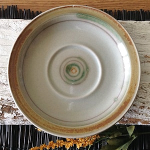 May include: A white ceramic saucer with a brown rim and a green and brown concentric circle design in the center.