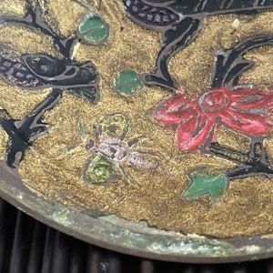 以下が含まれることがあります： 黒、赤、緑、白で描かれた花と葉が特徴的な、丸い金色の金属製のトレイ。トレイには、小さくピンクと緑の昆虫が描かれている。