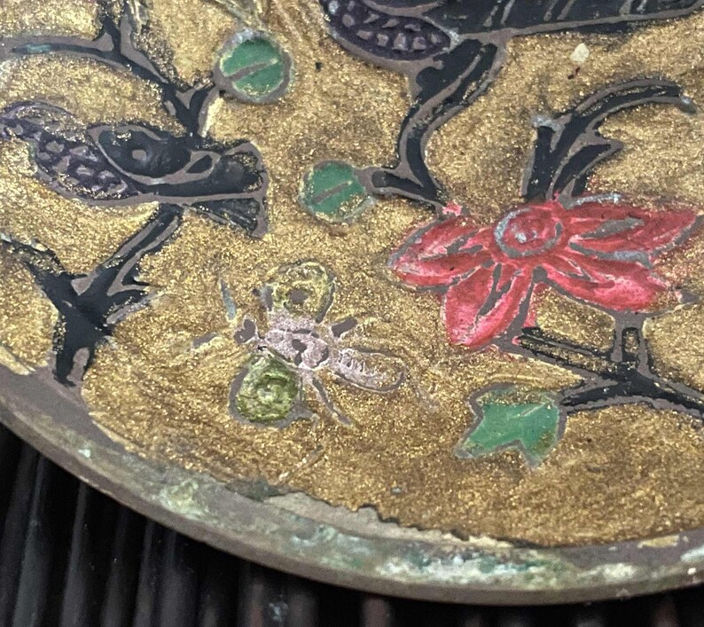 以下が含まれることがあります： 黒、赤、緑、白で描かれた花と葉が特徴的な、丸い金色の金属製のトレイ。トレイには、小さくピンクと緑の昆虫が描かれている。