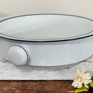 May include: White enamel oval baking dish with a dark blue and gray trim. The dish has a rounded handle on one side. The dish is sitting on a white and brown surface.