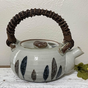 May include: A white ceramic teapot with a brown wicker handle and a leaf design. The teapot has a brown lid with a small knob.