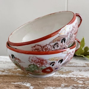 May include: Two stacked antique Japanese porcelain bowls. The bowls have a white interior and a red rim. The exterior features a detailed scene with figures, trees, and umbrellas in shades of red, brown, and gold.