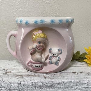 May include: A pink ceramic mug with a handle, featuring a raised relief design of a baby and a teddy bear inside a circular frame. The rim has blue floral accents, and the mug is inscribed with the word "Hers".