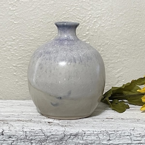 May include: A small, round, ceramic vase with a light gray and purple glaze. The vase has a slightly uneven surface and a rounded bottom.