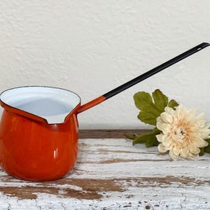 May include: A small, orange enamel saucepan with a long, black handle. The interior is white, and the handle has an orange accent near the pot. A cream-colored flower and green leaves sit nearby on a distressed white surface.