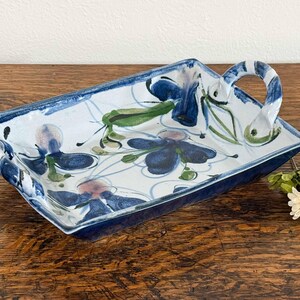 May include: A rectangular ceramic serving tray with two arched handles, featuring a white base with hand-painted dark blue floral motifs and green leaf accents. The edges are finished in a deep blue glaze, and the interior surface displays a pattern of blue flowers with thin, swirling blue lines.