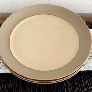 May include: A stack of three ceramic plates. Each plate has a light beige center and a wide, muted gray border. The plates are stacked on top of each other, showcasing their simple, modern design. The plates are suitable for everyday use.