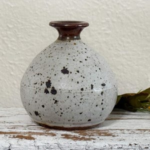 May include: A speckled ceramic vase with a rounded body and a dark brown rim. The vase has a light gray base color with dark brown speckles. The vase is a decorative item.