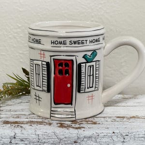 May include: White ceramic mug with a black and white illustration of a house with a red door and the text "Home Sweet Home".