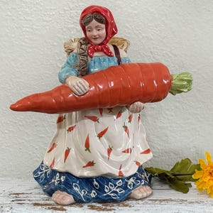 May include: A ceramic figurine of a woman in a red headscarf and blue dress, holding a large orange carrot. The woman's apron is white with red chili pepper designs. A yellow flower rests on the surface.