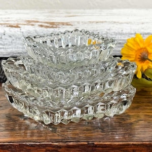 May include: A stack of three clear, square-shaped glass dishes with a textured, geometric pattern. The dishes are nested together, showcasing their varying sizes. A yellow flower with green leaves is in the background.